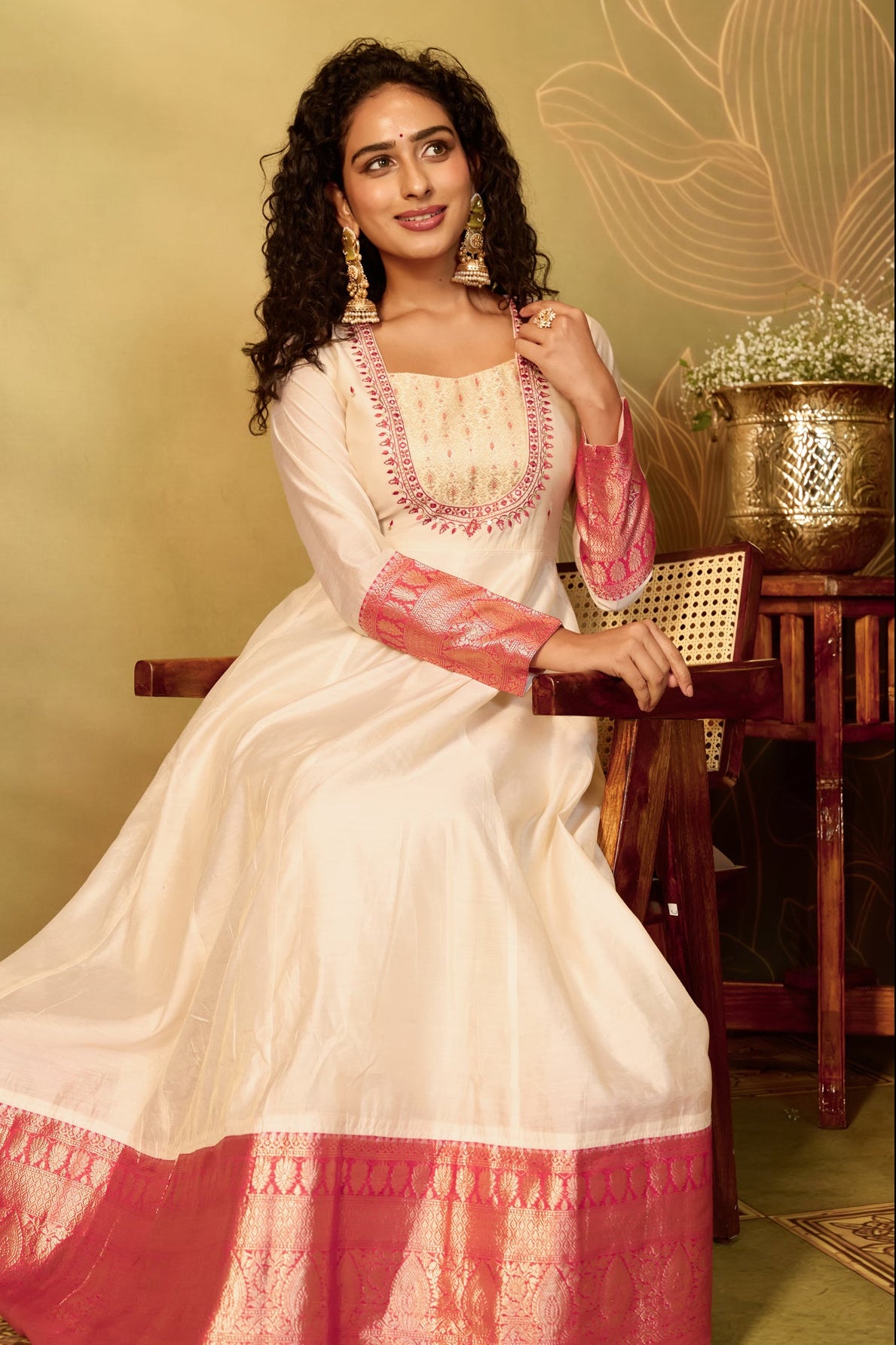 A woman models a traditional Indian Anarkali gown in off-white with vibrant pink and gold zari embroidery and borders.