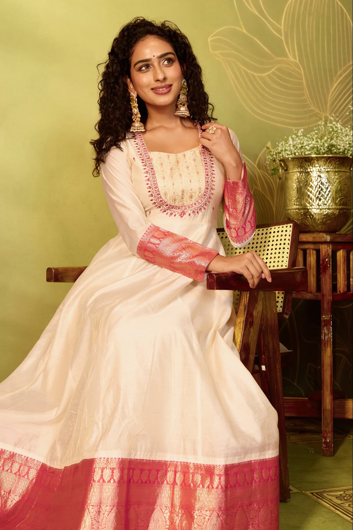 A woman wearing a traditional Festive Indian Anarkali gown in off-white with red and gold zari embroidery.